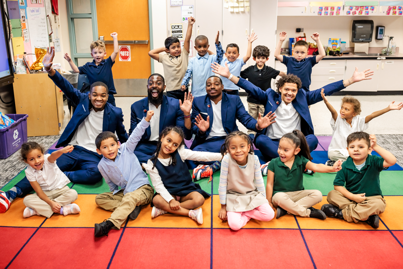 The MSU Denver Call Me MISTER students and director sit with a group of elementary school students on a colorful floor mat, arms extended and smiling.