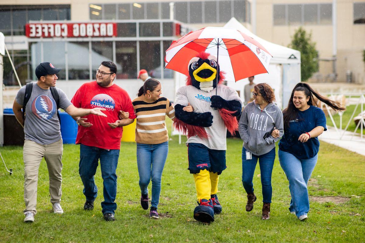 Five students walk in the rain with their arms linked with Rowdy the mascot in the middle holding a red and white striped umbrella